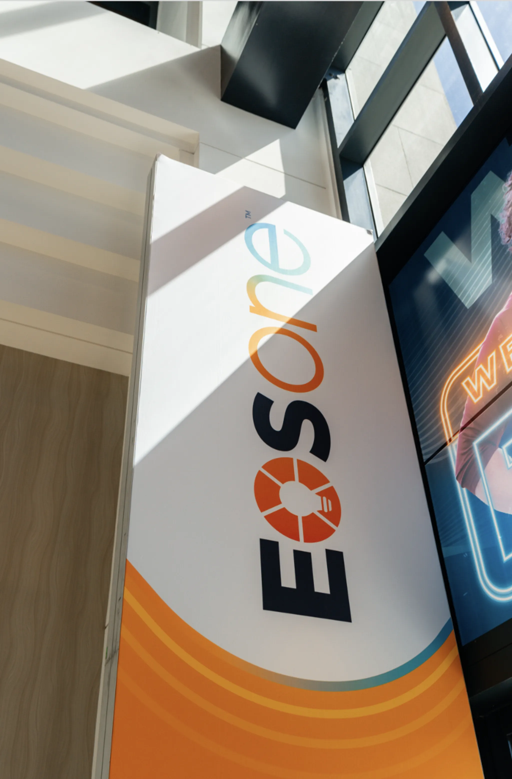 Tall EOS One™ branded standee in a conference foyer alongside an event schedule monitor
