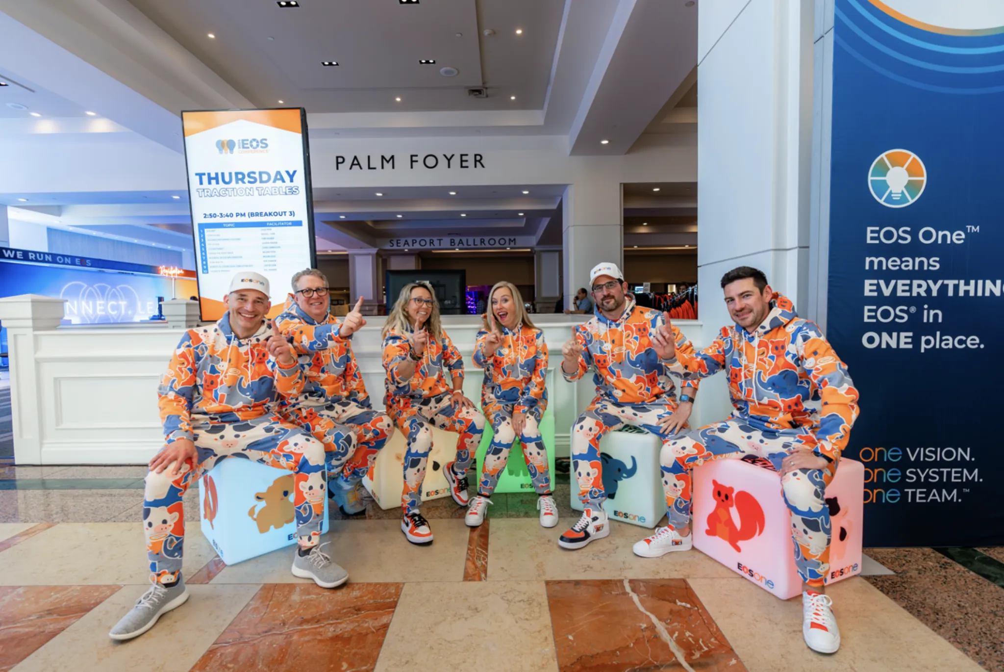 Six attendees in matching EOS-patterned suits seated on branded pedestals in front of an EOS One™ activation in the Palm Foyer