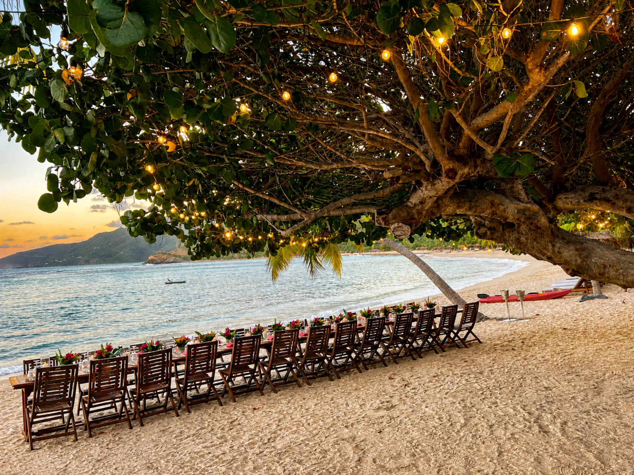 Long beachside dinner table set under a tree strung with lights at sunset with kayaks and Caribbean bay in the background