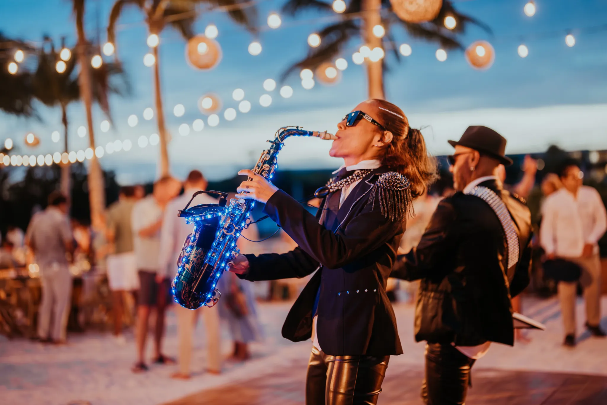 Saxophonist in a sequined blazer performing under string lights at a Seismic outdoor evening event.