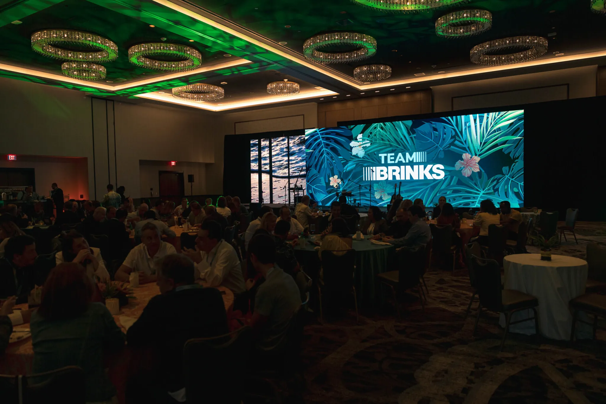 Attendees seated at round tables in a ballroom facing a large 'Team Brinks' tropical-themed stage screen.